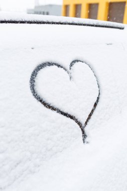 Kışın karla kaplı bir araba caddede durur. Arabaların camlarında kar var. Arabanın camında, karda boyanmış kalp.
