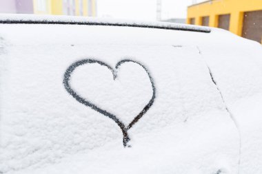 Kışın karla kaplı bir araba caddede durur. Arabaların camlarında kar var. Arabanın camında, karda boyanmış kalp.