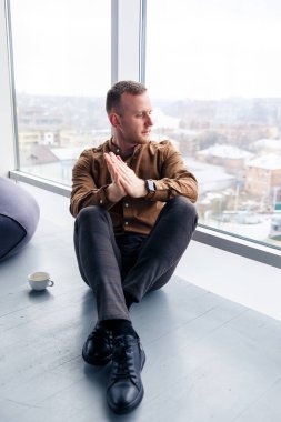 Yakışıklı genç erkek yönetici panoramik pencerenin yanında telefonla yerde oturuyor. Erkek işadamı yeni bir proje üzerinde çalışıyor.