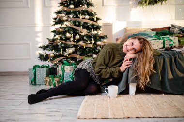 Sarışın, güzel genç bir kadın elinde kahveyle bir Noel ağacının yanında oturuyor. Yeni yıl yaklaşıyor. Sıcak bir evde Noel atmosferi