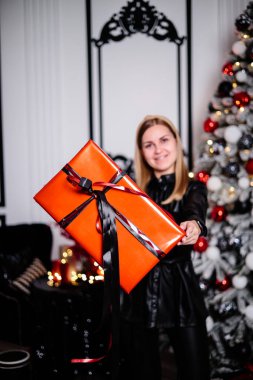 Güzel bir kadın çelenkle süslenmiş bir Noel ağacının yanında duruyor ve bir hediye açıyor. Yanındaki Noel ağacı hediye kutularıyla süslenmiş. Yeni Yıl