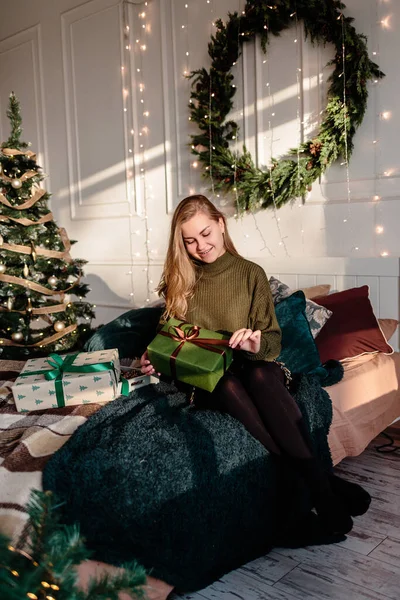 一位身穿毛衣的年轻女子在圣诞树旁装饰着圣诞装饰品的卧室里打开圣诞礼物 — 图库照片