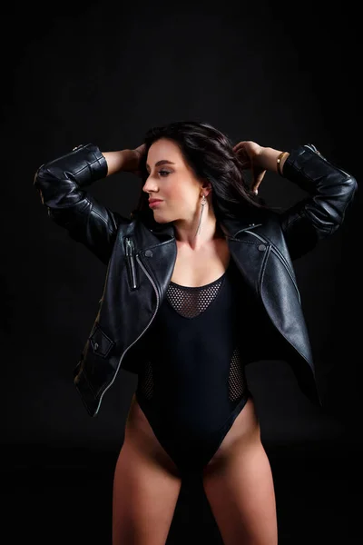 黒のボディスーツと黒の革のバイカージャケットで完璧な体を持つ細い日焼けした女性が黒の背景にスタジオでポーズ — ストック写真