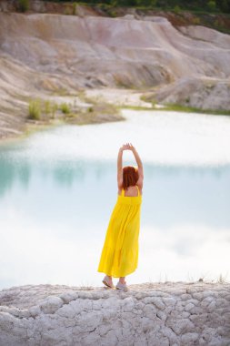 Göl kenarında sarı elbiseli genç bir kadın. Gök mavisi suyu ve yeşil ağaçları var..
