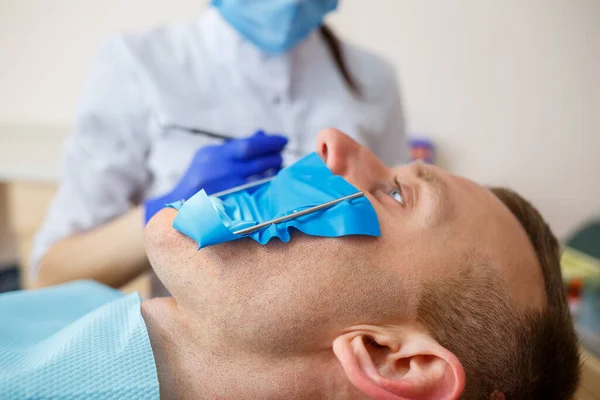 Жінка-стоматолог оглядає пацієнта за допомогою інструментів у стоматологічній клініці. Лікар робить стоматологічне лікування на зубах людини в стоматологічному кріслі. Вибірковий фокус — стокове фото