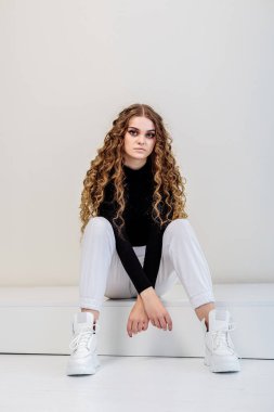Uzun kıvırcık saçlı, siyah kazaklı, beyaz pantolonlu, beyaz deri ayakkabılı güzel bir kadın. Yeni kadın ayakkabıları koleksiyonu.
