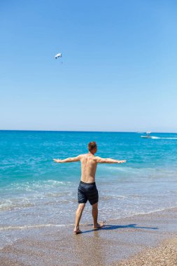 Güzel vücutlu çekici bir genç adam kumlu sahil boyunca yürüyor ve yaz tatilinin tadını çıkarıyor.