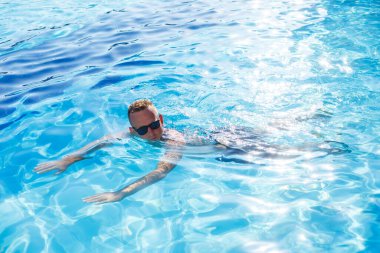 Young attractive man in sunglasses is resting in the pool on summer vacation. The guy in the pool by the hotel