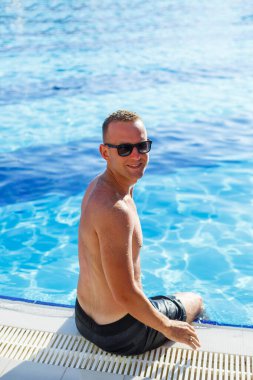 Young attractive man in sunglasses is resting in the pool on summer vacation. The guy in the pool by the hotel