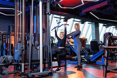 Deneyimli bir özel antrenör, son teknoloji spor salonunda kas egzersizleri sırasında atletik bir kadını denetliyor..