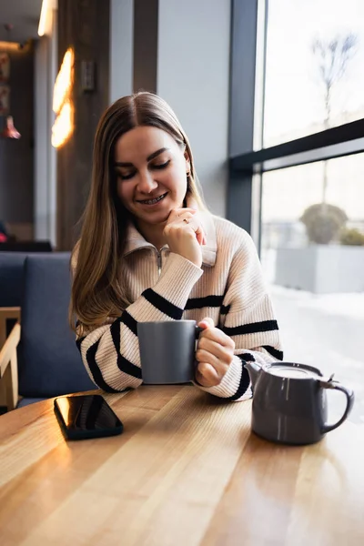 坐在餐馆的餐桌前 一个漂亮而沉着的年轻女子一边喝咖啡 一边微笑着看着窗外 晨咖啡 — 图库照片