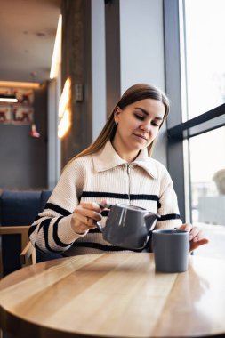 Güzel bir kadın bir fincana çay dolduruyor, pencerenin yanındaki bir masada oturuyor. Genç bayan sabahtan zevk alıyor. Sabah sıcak aromalı çay.