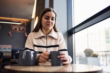 Güzel, gülümseyen, sakin, kahve içen genç bir kadın bir restoranda masada otururken pencereden dışarı bakıyor. Sabah kahvesi