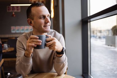 Pozitif bir adam bir kafede oturur ve aromatik kahve içer. Günlük kıyafetlerle gülümseyen adam bir kafede oturur ve sabah kahvesini içer.
