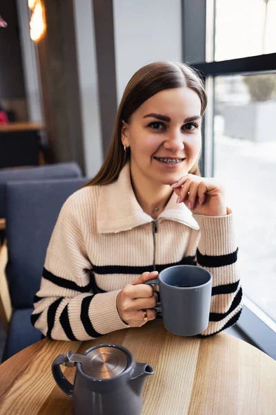 坐在餐馆的餐桌前 一个漂亮而沉着的年轻女子一边喝咖啡 一边微笑着看着窗外 晨咖啡 — 图库照片