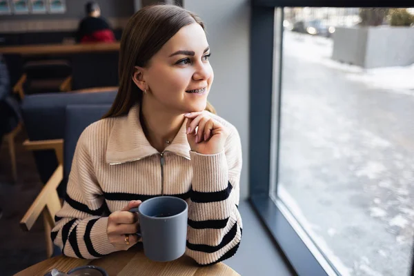 一位年轻漂亮的女士坐在窗边一张桌子旁的咖啡店里 一边喝咖啡 一边朝窗外看 年轻的女人正在享受早晨的快乐 早晨一杯令人神清气爽的咖啡 — 图库照片