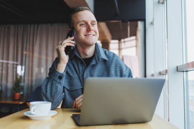 Gündelik giysiler içinde gülümseyen erkek girişimci dizüstü bilgisayarla masada oturuyor ve modern çalışma alanında gün içinde geniş pencereli akıllı telefonla konuşuyor.