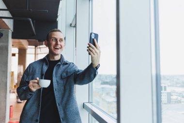 Takım elbiseli genç adam cep telefonunu komik bir ifadeyle kullanıyor. Akıllı telefonuna bakıp iyi haberleri e-posta yoluyla okuyor. Duygusal yaşam tarzı kavramı..