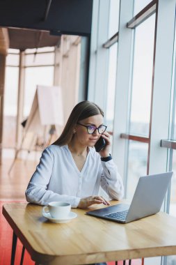 Web dosyaları oluşturmak için kablosuz internete bağlanan grafiksel tasarımcı, fikirleri tartışmak için meslektaşını arayan, gözlüklü gülümseyen kadın, akıllı telefonla konuşuyor ve kafede kahve içiyor