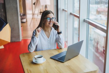 Genç ve güzel bir kadın kafede oturmuş konuşurken gülüyor, sipariş veriyor ve dizüstü bilgisayarına not alıyor. Kahve içerken cep telefonundan arayan pozitif kız.