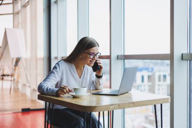 Genç ve güzel bir kadın kafede oturmuş konuşurken gülüyor, sipariş veriyor ve dizüstü bilgisayarına not alıyor. Kahve içerken cep telefonundan arayan pozitif kız.
