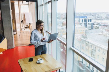 Beyaz gömlekli, gözlüklü, Avrupalı bir iş kadını elinde taşınabilir bir laptop, en üst kattaki ofisin büyük bir penceresinin yanında duruyor.