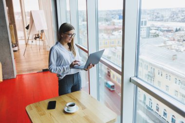 Beyaz gömlekli, gözlüklü, Avrupalı bir iş kadını elinde taşınabilir bir laptop, en üst kattaki ofisin büyük bir penceresinin yanında duruyor.