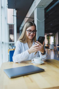 Mutlu gülümseyen bayan yönetici internet uygulaması üzerinden dağıtım hizmetlerini kontrol etmek için cep telefonu aygıtı kullanıyor, kahve dükkanında ücretsiz 4G kablosuz ağ, cep telefonuyla mesajlaşan neşeli serbest çalışan
