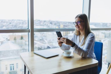 Mutlu gülümseyen bayan yönetici internet uygulaması üzerinden dağıtım hizmetlerini kontrol etmek için cep telefonu aygıtı kullanıyor, kahve dükkanında ücretsiz 4G kablosuz ağ, cep telefonuyla mesajlaşan neşeli serbest çalışan