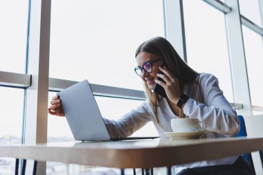 Web dosyaları oluşturmak için kablosuz internete bağlanan grafiksel tasarımcı, fikirleri tartışmak için meslektaşını arayan, gözlüklü gülümseyen kadın, akıllı telefonla konuşuyor ve kafede kahve içiyor