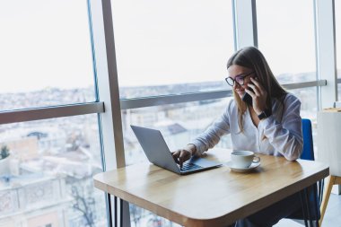 Web dosyaları oluşturmak için kablosuz internete bağlanan grafiksel tasarımcı, fikirleri tartışmak için meslektaşını arayan, gözlüklü gülümseyen kadın, akıllı telefonla konuşuyor ve kafede kahve içiyor