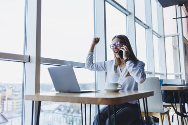 Web dosyaları oluşturmak için kablosuz internete bağlanan grafiksel tasarımcı, fikirleri tartışmak için meslektaşını arayan, gözlüklü gülümseyen kadın, akıllı telefonla konuşuyor ve kafede kahve içiyor