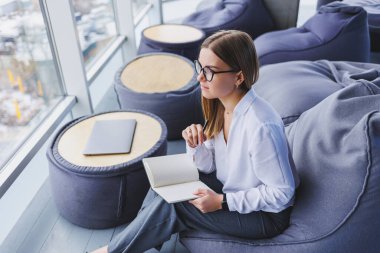 Ayrıl. Gözlüklü, gülümseyen genç bir kadın masada oturuyor ve rahatlıyor. Not defteri kullanıyor, mutlu bir kadın arkasını yaslıyor ve işten zevk alıyor.