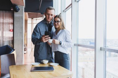 En üst kattaki büyük bir pencerenin yanında modern bir ofisin masasında dikilirken telefondaki verileri kontrol eden ve not alan sıradan giyinen zeki genç profesyoneller.