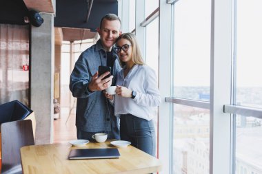 En üst kattaki büyük bir pencerenin yanında modern bir ofisin masasında dikilirken telefondaki verileri kontrol eden ve not alan sıradan giyinen zeki genç profesyoneller.