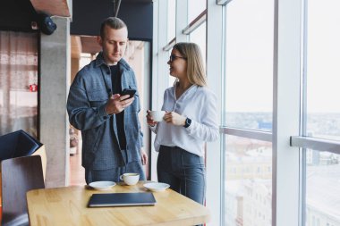 En üst kattaki büyük bir pencerenin yanında modern bir ofisin masasında dikilirken telefondaki verileri kontrol eden ve not alan sıradan giyinen zeki genç profesyoneller.