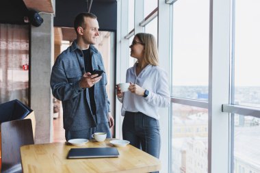 En üst kattaki büyük bir pencerenin yanında modern bir ofisin masasında dikilirken telefondaki verileri kontrol eden ve not alan sıradan giyinen zeki genç profesyoneller.