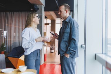 En üst kattaki büyük bir pencerenin yanında modern bir ofisin masasında dikilirken telefondaki verileri kontrol eden ve not alan sıradan giyinen zeki genç profesyoneller.