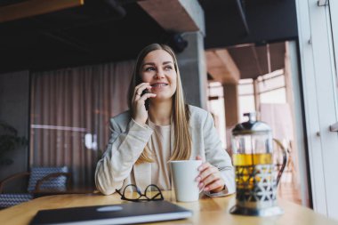 Telefon görüşmeleri sırasında iş problemlerini çözen başarılı genç bir kadın, modern bir kafede oturuyor ve bir dizüstü bilgisayarla çalışıyor. Hoş bir iç mekan, kafede boş vaktinin tadını çıkarıyor..