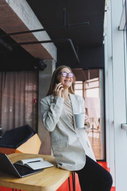 Telefon görüşmeleri sırasında iş problemlerini çözen başarılı genç bir kadın, modern bir kafede oturuyor ve bir dizüstü bilgisayarla çalışıyor. Hoş bir iç mekan, kafede boş vaktinin tadını çıkarıyor..