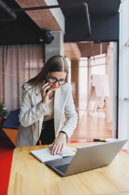 Telefon görüşmeleri sırasında iş problemlerini çözen başarılı genç bir kadın, modern bir kafede oturuyor ve bir dizüstü bilgisayarla çalışıyor. Hoş bir iç mekan, kafede boş vaktinin tadını çıkarıyor..