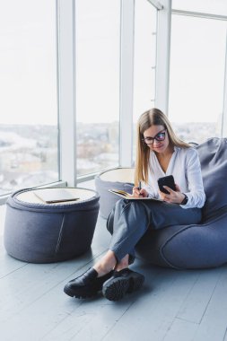 Kahve dükkanındaki güzel Avrupalı genç bir iş kadını cep telefonuyla oturur ve not defterine not alır. Şık bir takım elbise giyip halka açık bir kafede dizüstü bilgisayarla oturmak.
