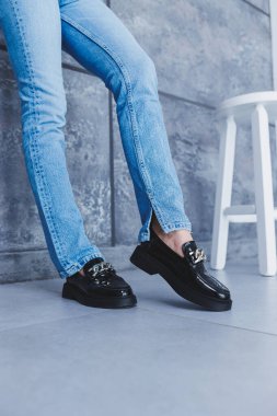 İnce, güzel bacakları ve şık siyah deri ayakkabıları olan genç bir kadın stüdyoda bir sandalyede oturuyor. Kadın mokasenleri, yakın plan. Bahar ayakkabısı koleksiyonu