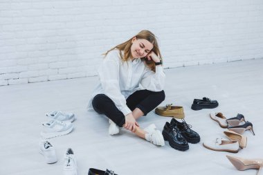 Genç bir kadın ayakkabılar arasında yerde oturuyor ve kendisi için yeni ayakkabılar seçiyor. Beyaz gömlekli ve siyah pantolonlu bir bayan. Beyaz ayakkabılı kadın..