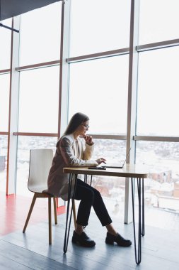 Modern bir ofiste geniş pencereli bir ofiste telefonla konuşurken ceketli, gülümseyen ve dizüstü bilgisayarla çalışan mutlu genç bir kadının görüntüsü. Uzak çalışma