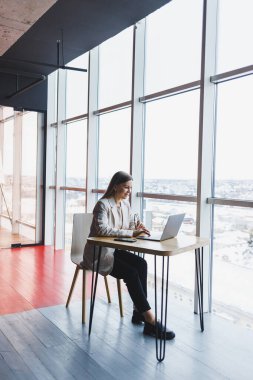 Modern bir ofiste geniş pencereli bir ofiste telefonla konuşurken ceketli, gülümseyen ve dizüstü bilgisayarla çalışan mutlu genç bir kadının görüntüsü. Uzak çalışma