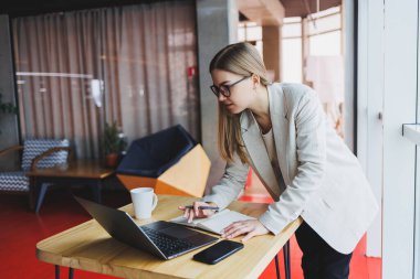 Ciddi genç iş kadını ahşap bir masada oturuyor ve modern bir çalışma alanında dizüstü bilgisayar üzerinde çalışırken kalemle notlar yazıyor. Gözlüklü ve ceketli kız serbest çalışıyor.