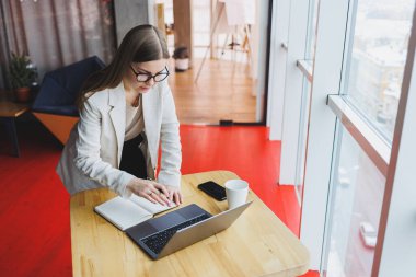 Ciddi genç iş kadını ahşap bir masada oturuyor ve modern bir çalışma alanında dizüstü bilgisayar üzerinde çalışırken kalemle notlar yazıyor. Gözlüklü ve ceketli kız serbest çalışıyor.