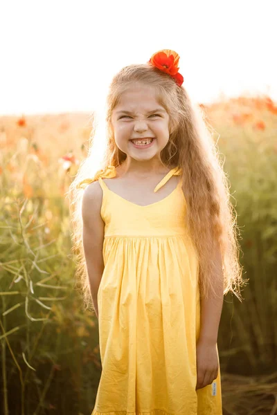 畑のポピーの中で彼女の顔に笑顔で陽気な女の子 赤い花 黄色のショートドレスと帽子のブロンド — ストック写真