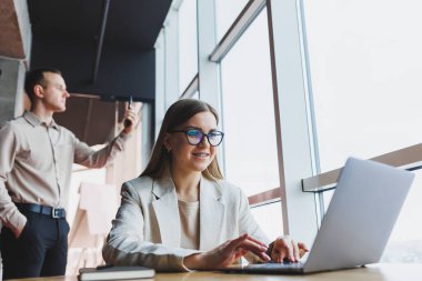 Ceketli neşeli bir bayan yönetici ofis işi yapıyor ve masasında poz vermek için optik gözlüklü başarılı bir bayan patron. Arka planda telefonu olan bir adam var.